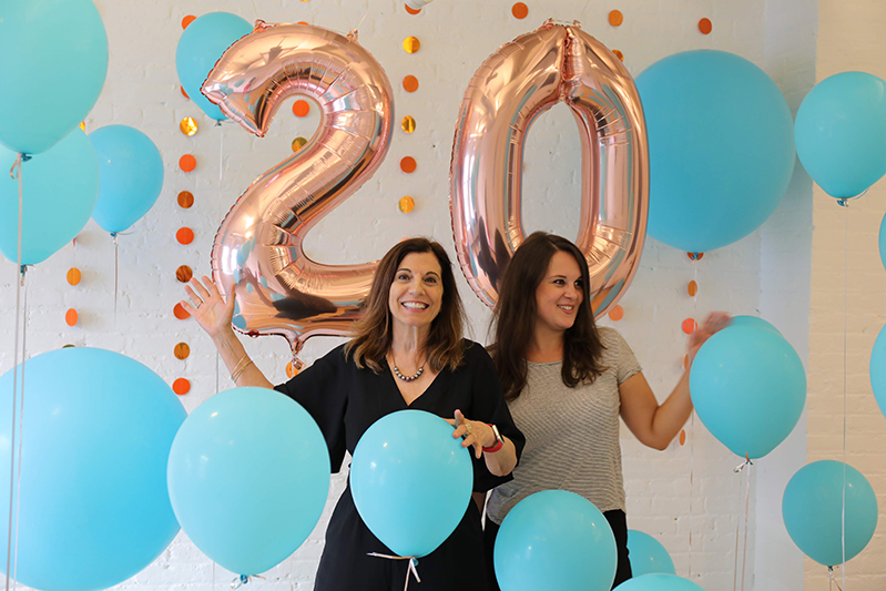 A photo of two women surrounded by blue balloons and in the background the number twenty made also from shiny balloons.