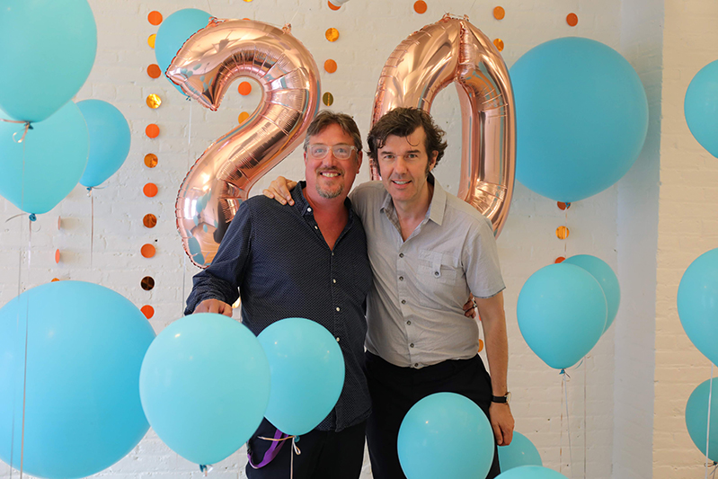 A photo of two men surrounded by blue balloons and in the background the number twenty made also from shiny balloons.