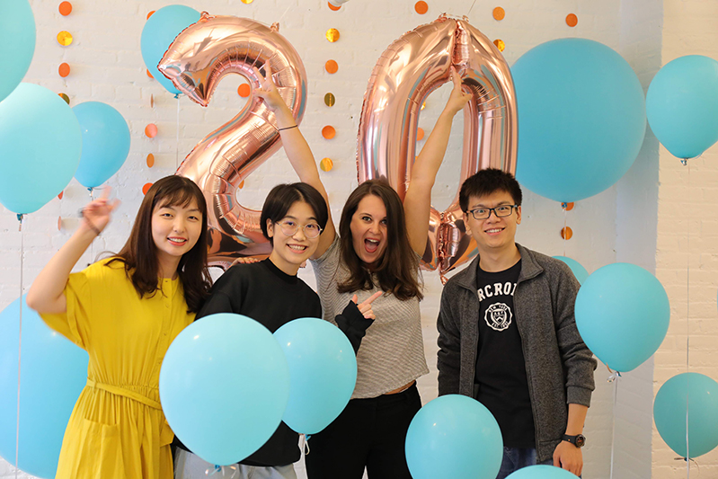 A photo of a group of students surrounded by blue balloons and in the background the number twenty made also from shiny balloons.