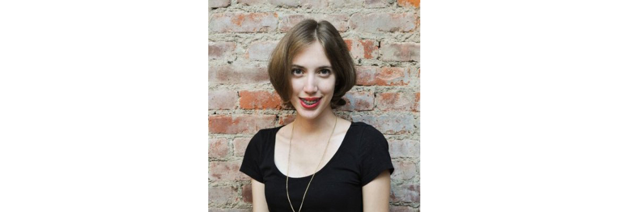 A photo of a woman wearing a black t-shirt and standing in front of a brick wall.