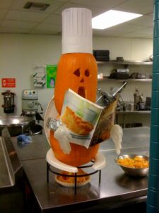 A photo of a pumpkin head wearing a chef's head, hands made from chirurgical white gloves and a pumpkin body. The figure reads a food magazine while sitting on a made up toilet seat. Some leftovers are underneath and beside it in a bowl.