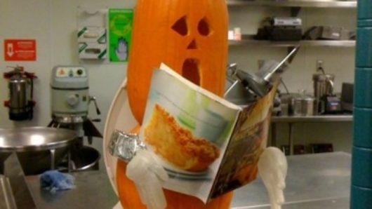 A photo of a pumpkin head wearing a chef's head, hands made from chirurgical white gloves and a pumpkin body. The figure reads a food magazine while sitting on a made up toilet seat. Some leftovers are underneath and beside it in a bowl.