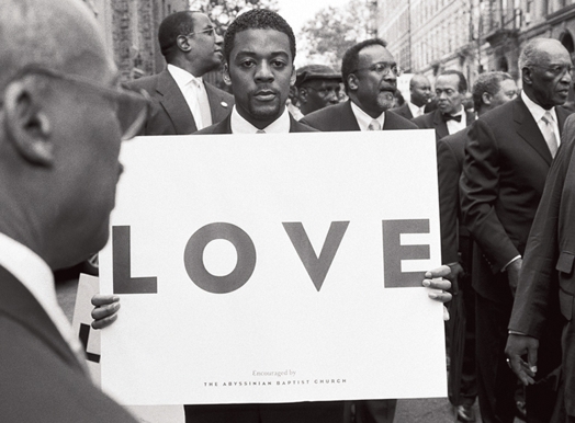 A black and white photo of a man in a suit holding a placard with the word LOVE while standing among other people wearing suits.