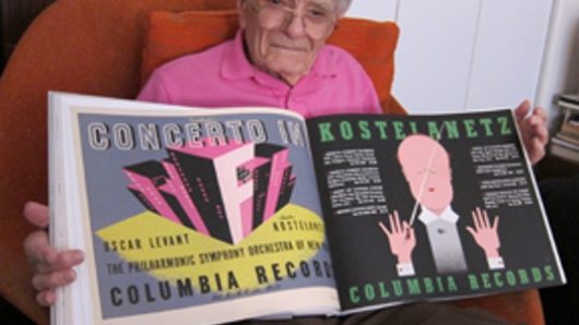 A photo of a man with glasses and pink shirt, sitting in an armchair and holding a design book containing some of his work.