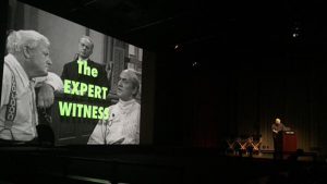 A photo showing a man giving a lecture while on a projector screen near him is a black and white photo with the green text The Expert Witness above it.