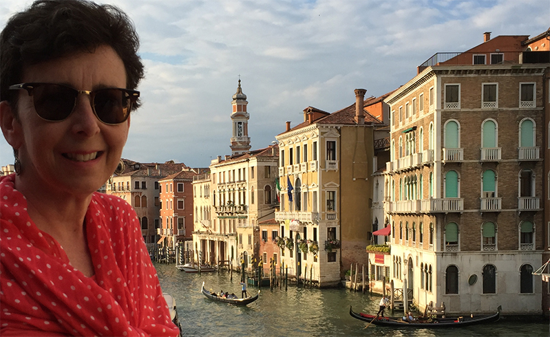 A photo of a woman wearing glasses and a red dress while in the background the cityscape of Venice.