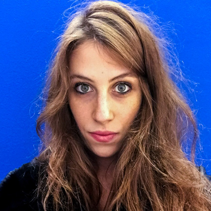 A photo of a woman wearing a black blouse and setting in front of a blue background.