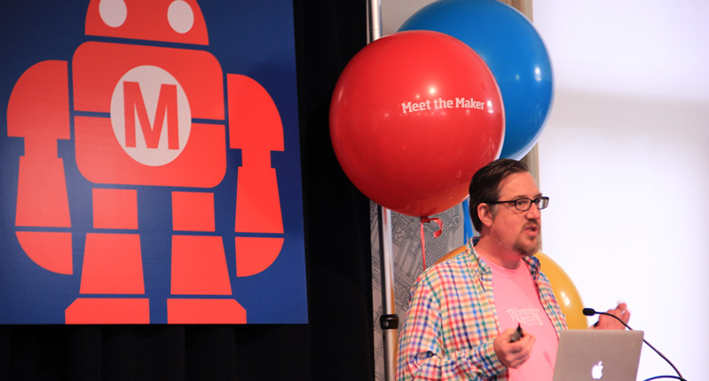 A photo of a person giving a lecture while sitting in front of a panel on which a red robot with an M is drawn. Also there are a blue and red balloons with text Meet The Makers on them.