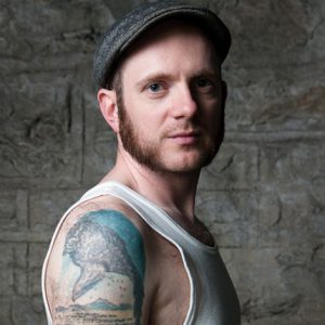 A photo of a tattooed man wearing a white undershirt and a hat.