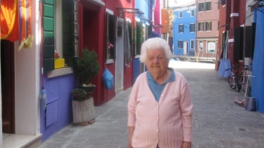 A photo of an old woman caring a bag while sitting in a narrow paved street near some colorful houses.