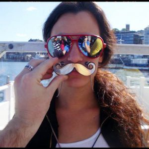 A photo of a woman wearing sunglasses while a man holds a fake metal moustache against her face.