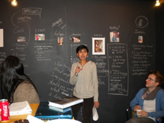 A photo of a person giving a lecture and sitting in front of a blackboard like wall with chalk writings and pictures on it.