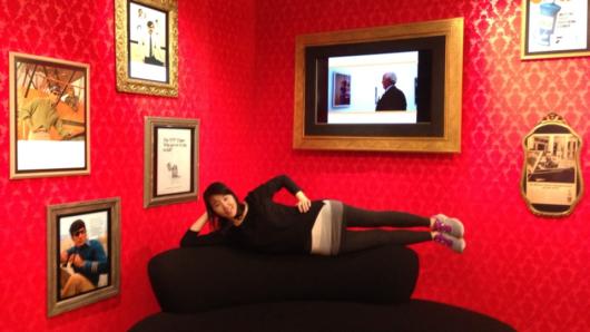 A photo of a girl sitting on a couch and posing. The couch is placed in a room with red textured walls and different photographs.