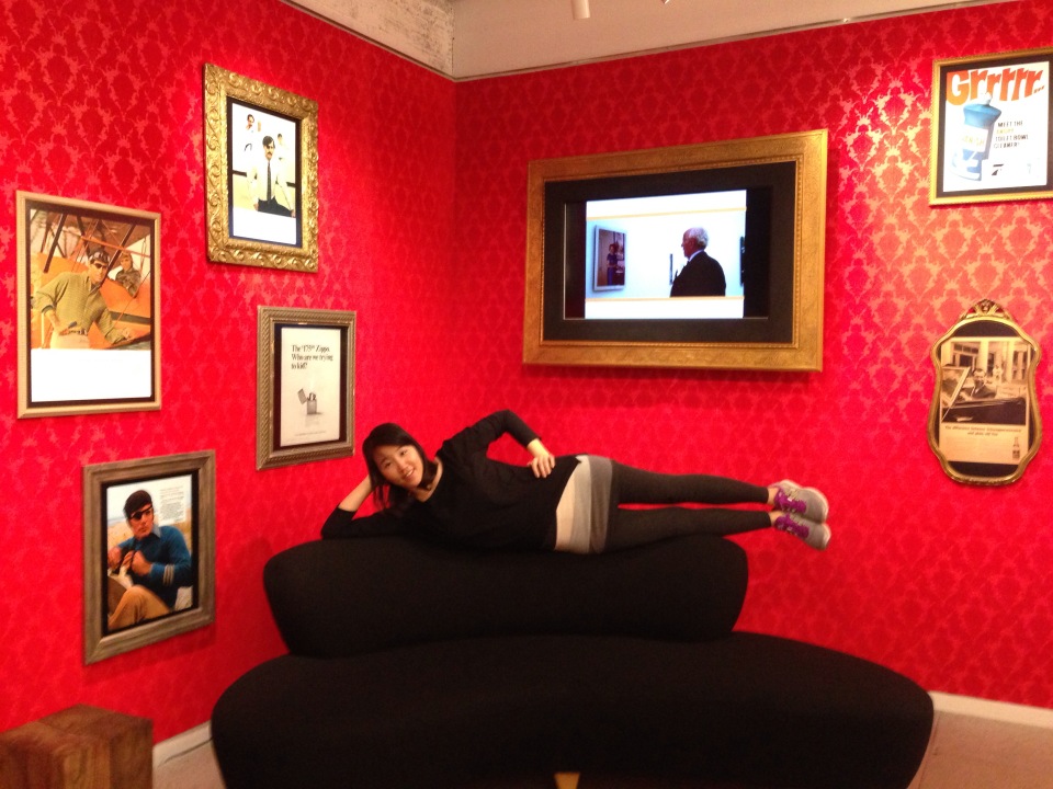 A photo of a girl sitting on a couch and posing. The couch is placed in a room with red textured walls and different photographs.