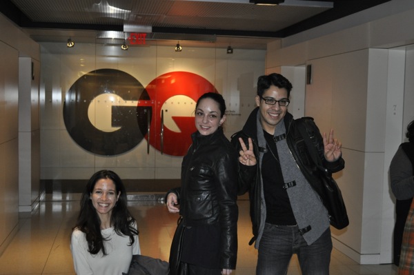 A photo of three persons standing in a room in front of a logo commercial with letters GQ.