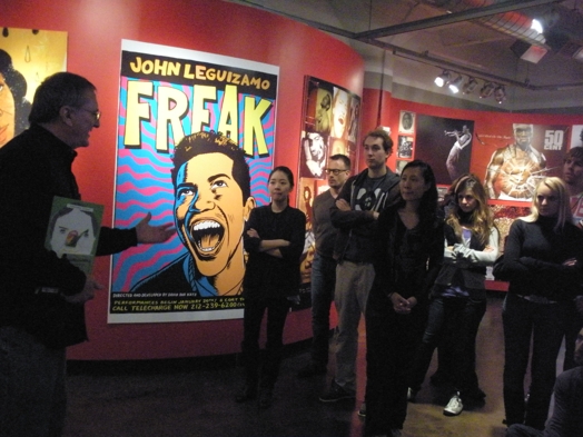A group of people listening to a man while sitting in an exhibition gallery near a cartoon poster.