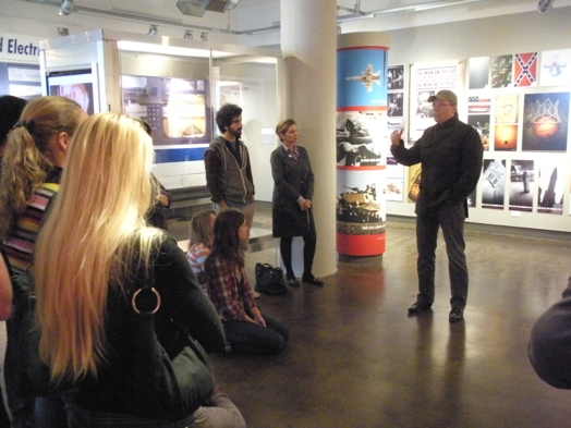 A photo of a group of people listening to a spokesman while sitting in an exhibition gallery.