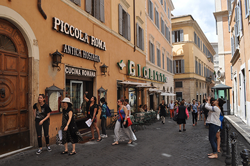 A photo of a paved street in Rome filled with people walking by some buildings.