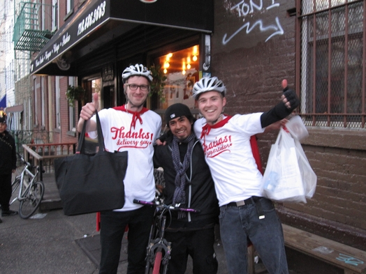 A photo o three people, one on a bicycle and the other two wearing biking helmets and white t-shirts with a company logo. They are also holding bags in their hands while sitting in the street behind a restaurant entrance.