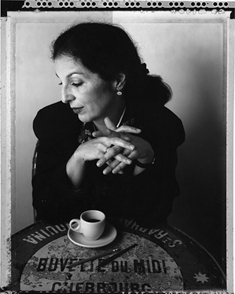 A black and white photo of a woman sitting and drinking coffee at a table with some words written on it.