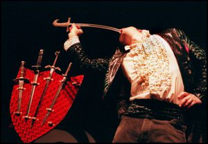 A photo of a man swallowing a sword and near him a shield with multiple swords on it