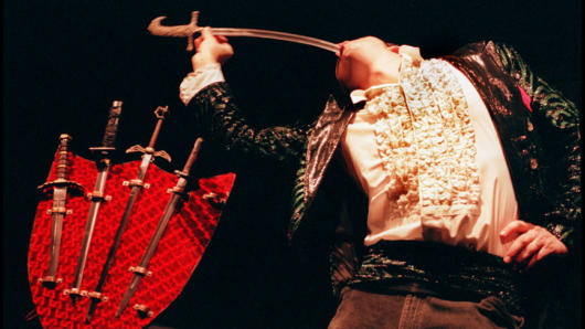 A photo of a man swallowing a sword and near him a shield with multiple swords on it