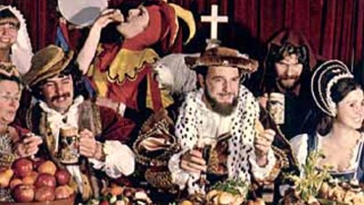An old photo of some medieval people eating at a table filled with food.