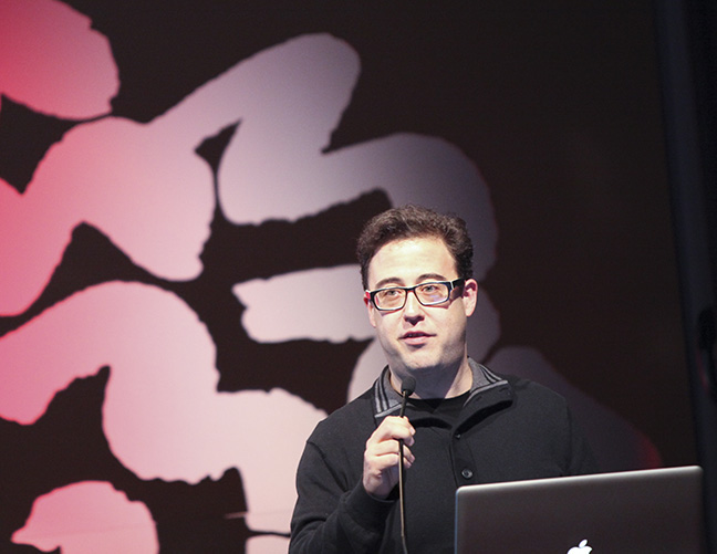 A photo of a man wearing glasses and talking on the microphone.