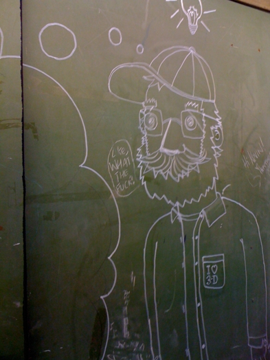A photo of a blackboard that has a chalk drawing of a man wearing glasses, a cap, a shirt and a moustache.