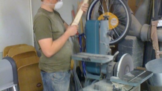 A photo of a man working on a wooden guitar handle in a woodworking workshop.