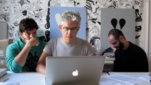 A photo of three people working at a laptop in an art workshop filled with canvases of drawn cartoons.