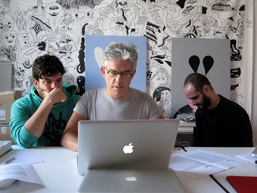 A photo of three people working at a laptop in an art workshop filled with canvases of drawn cartoons.