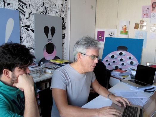 A photo of two people working at a laptop in an art workshop filled with canvases of drawn cartoons.