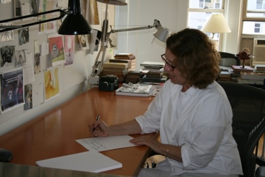 A photo of a woman wearing glasses and writing something on a peace of paper while standing at a desk.