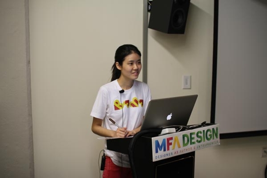 A photo of a woman giving a lecture from a laptop put on a stand with text MFA Design on it.