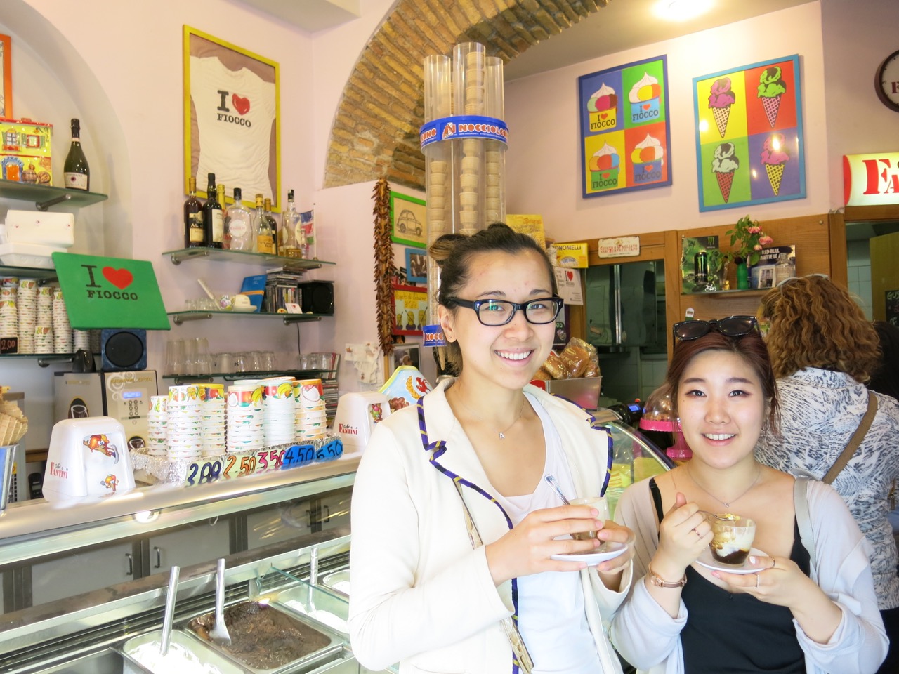 A photo of two women holding some plates with coffee cups while standing in a cafeteria.