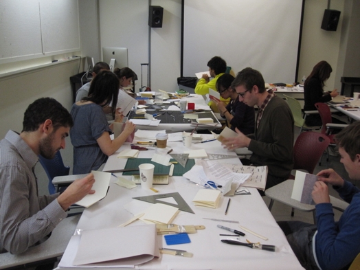 A photo of a group of students working with paper, trying to make a custom invitation card while sitting at a table filled with office items.