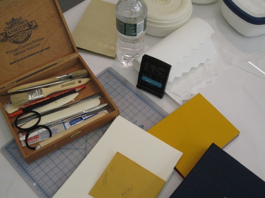A photo of different office items used when working with paper: a wooden box filled with brushes, pencils, tweezers, glue, crayons and rubber bands, several pieces of paper and cardboard, a cutting mat, a round plastic box and a bottle of water.