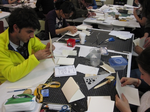 A photo of a group of students learning how to fold paper using different office items.