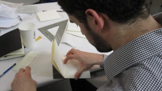 A photo of a man folding a piece of paper while sitting on a table filled with papers, a pencil, a square ruler, a notebook and a disposable coffee cup.