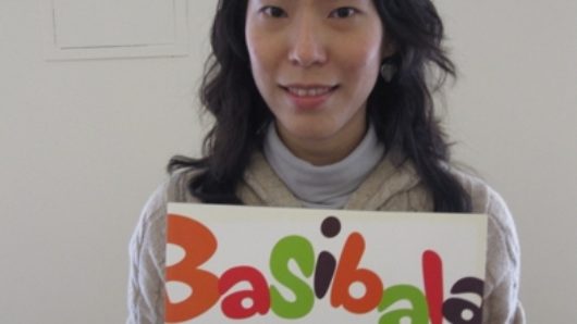 A photo of a women holding a banner with colorful letter text that says: Basibala.