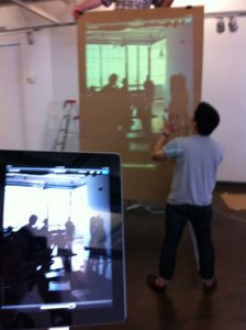 A photo of a tablet screen and a man standing and lookin at a projector screen held by another man.