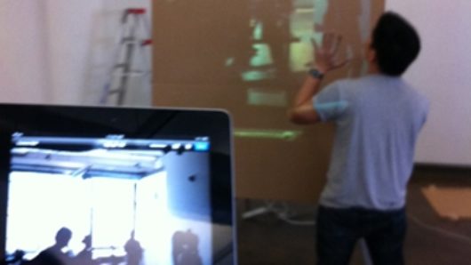 A photo of a tablet screen and a man standing and lookin at a projector screen held by another man.