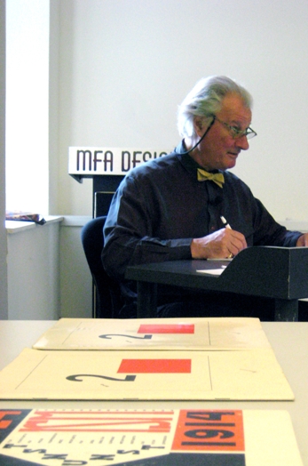 A photo of a man writing something on a piece of paper while sitting at a desk. Also some old styled magazines are on display near him.