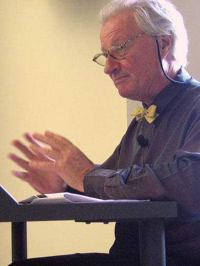 A photo of a man with glasses giving a lecture, while sitting at a desk with his papers.