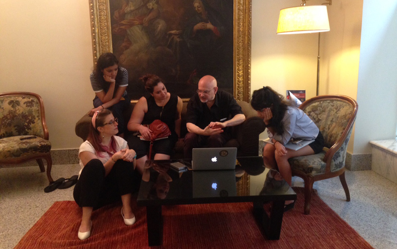 A photo of a group of people sitting in a room and talking to each other while looking at a laptop. Also behind them there is an old painting.