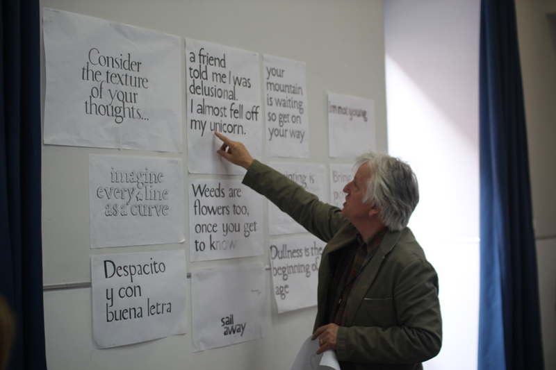 A photo of a teacher showing some pieces of paper with text on a wall and talking about different calligraphy stiles.