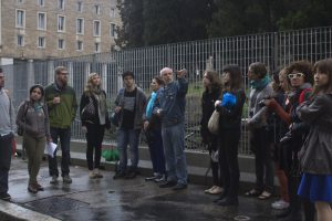 A photo of a group of people standing in the street and listening to a person giving a lecture and pointing at different buildings.