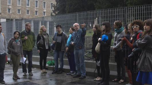 A photo of a group of people standing in the street and listening to a person giving a lecture and pointing at different buildings.