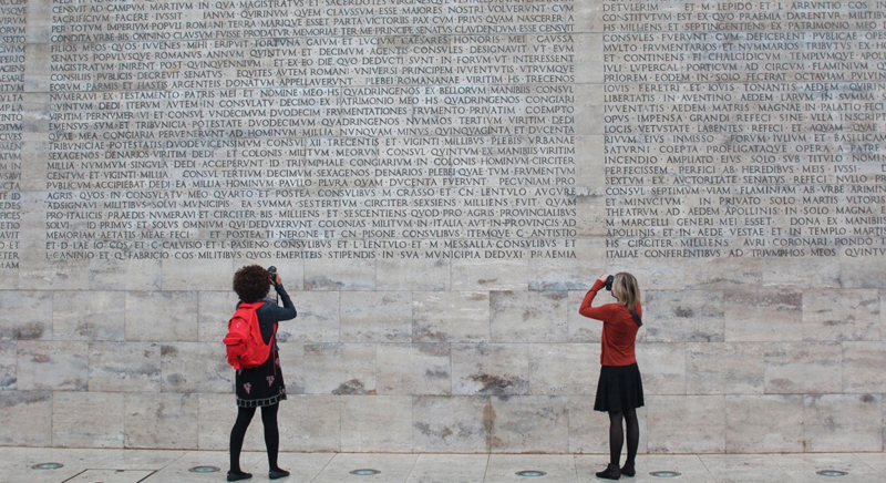 A photo of two students taking pictures of a stone wall engraved with text.